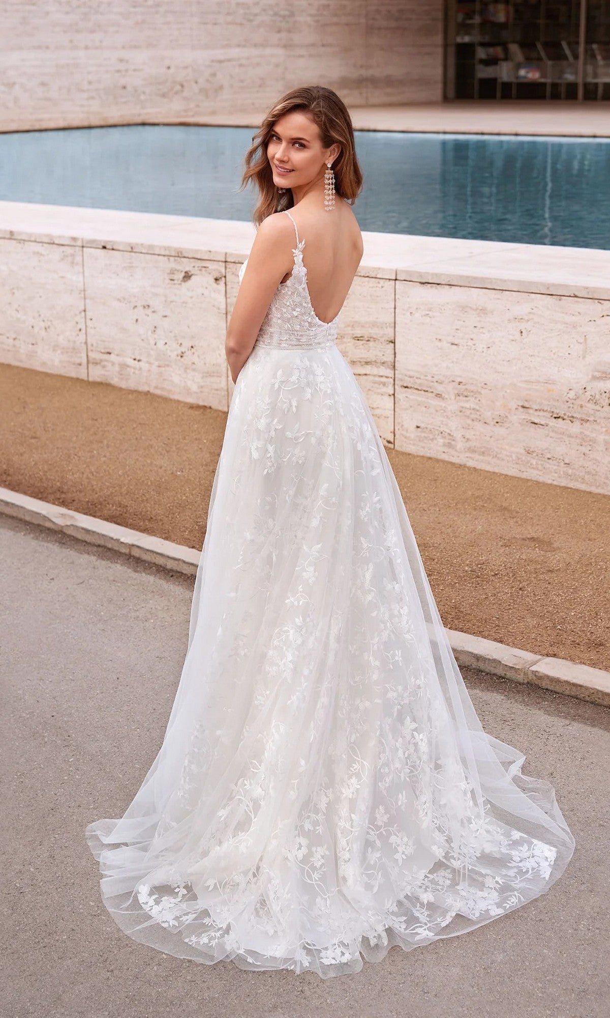 A woman in the Long White Dress: Alyce 7036, featuring floral appliqués, stands outdoors by a pool and gazes over her shoulder.